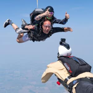 Tandem skydiving pair in freefall above open farmland, smiling and giving thumbs up as another skydiver captures the jump on camera.