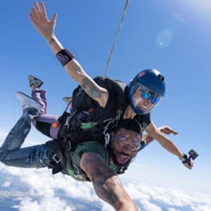 Tandem skydiving pair in freefall above the clouds on a clear day, with the instructor smiling and the passenger reaching toward the camera.