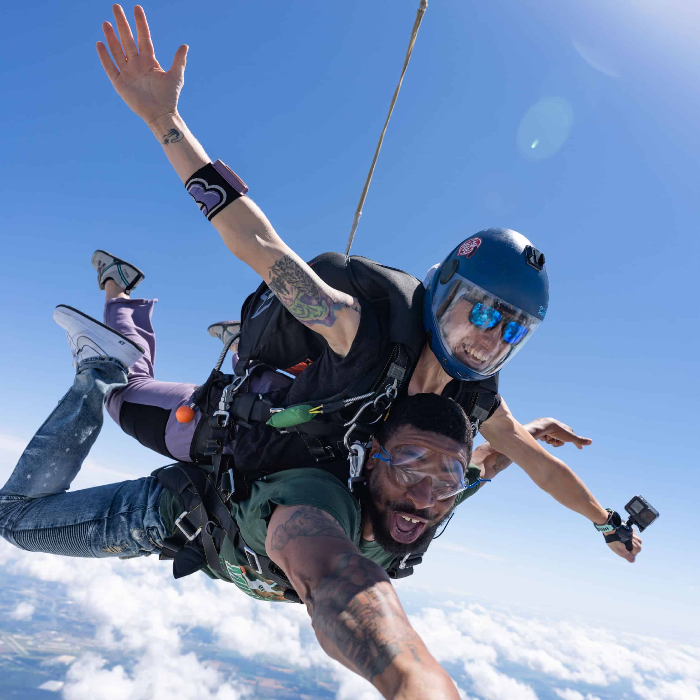 Tandem skydiving pair in freefall above the clouds on a clear day, with the instructor smiling and the passenger reaching toward the camera.